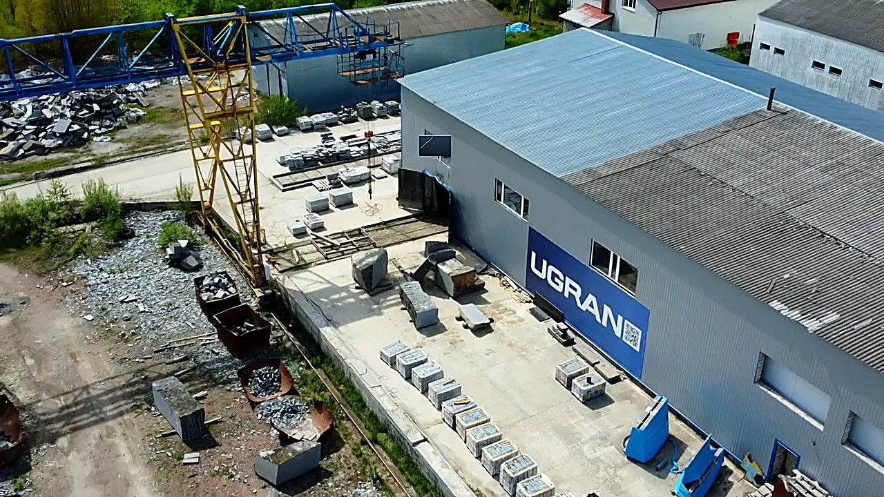 Производственная площадка UGRAN: корпус с вывеской, кран-балка, паллеты камня на бетонной площадке
